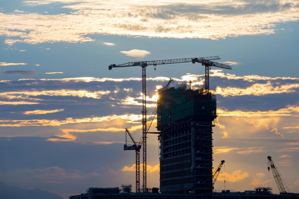 Silhouette of cranes and a building under construction against a vivid sunset sky.